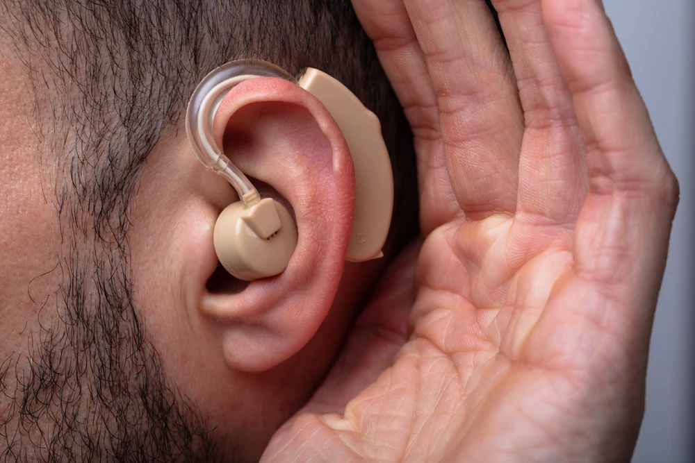Un homme porte un appareil auditif beige positionné sur son oreille droite, sa main gauche formant un geste d'écoute près de l'oreille