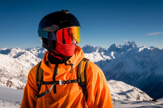 Skieur en haute montagne portant une veste orange technique, casque noir et masque de ski à verres miroir rouge-orange avec protection faciale
