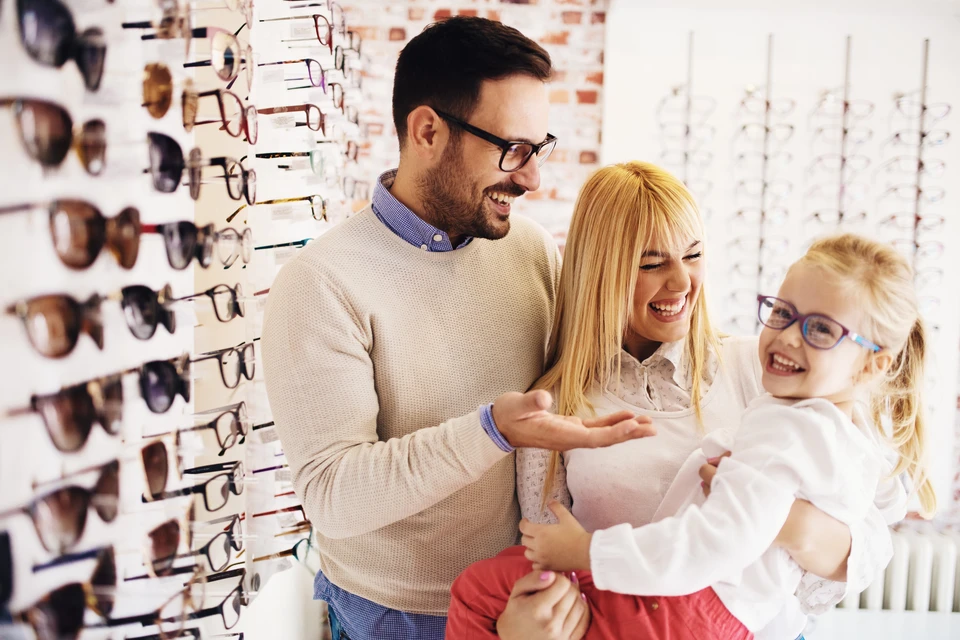 Famille souriante dans un magasin d'optique, père, mère et fillette blonde avec lunettes, consultant l'offre de montures
