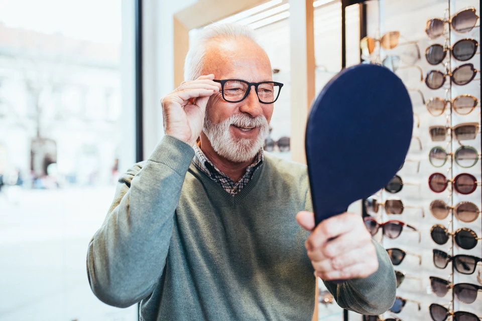 Homme âgé souriant dans un magasin d'optique, tenant un miroir pour essayer des lunettes de vue à monture noire