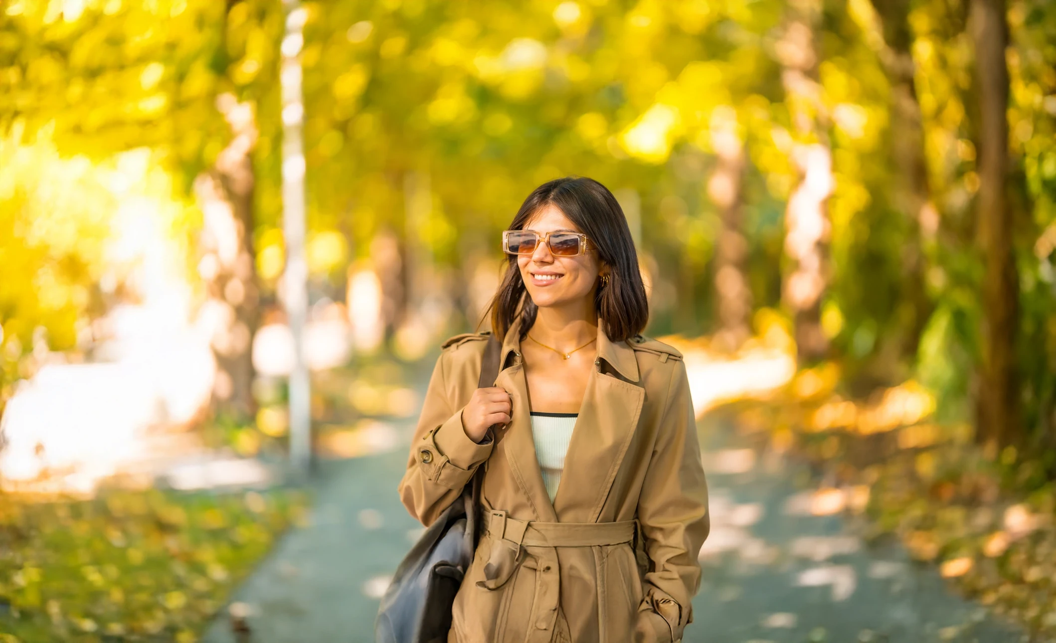 Jeune femme blonde aux cheveux ondulés porte des lunettes de soleil rondes dorées avec verres bleus foncés, écouteurs blancs, vêtement blanc, en extérieur avec arrière-plan flou naturel